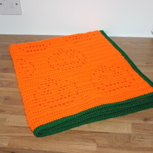 May include: An orange crochet blanket with a green border is folded on a wooden surface. The blanket is made of a textured yarn and has a geometric pattern. The green border provides a contrasting accent to the bright orange color.