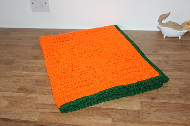 May include: An orange crochet blanket with a green border is folded on a wooden surface. The blanket is made of a textured yarn and has a geometric pattern. The green border provides a contrasting accent to the bright orange color.