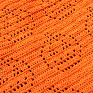 May include: Close-up of an orange crochet fabric with a diagonal pattern of openwork squares. The texture is visible, showing the individual stitches and the overall design. The bright color and open weave suggest a decorative item.