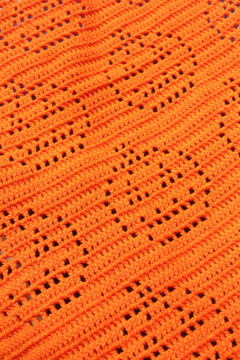 May include: Close-up of an orange crochet fabric with a diagonal pattern of openwork squares. The texture is visible, showing the individual stitches and the overall design. The bright color and open weave suggest a decorative item.