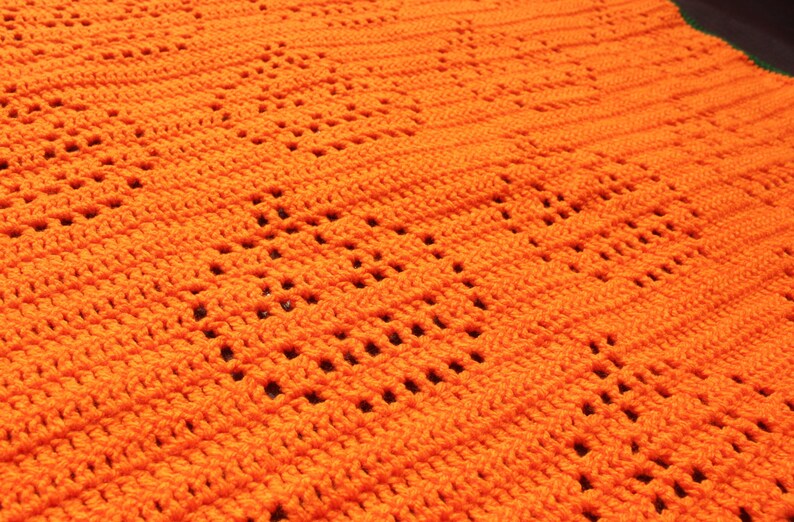May include: Close-up of a handmade orange crochet blanket. The blanket features a repeating pattern of openwork squares, creating a textured surface. The bright orange color and intricate design suggest a cozy, handcrafted item, perfect for home decor.
