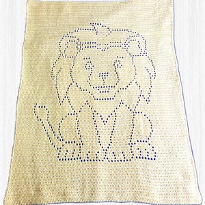 May include: A rectangular cream-coloured crochet blanket featuring a dark blue lion graphic created with a filet crochet technique. The lion design is centred on the blanket, showcasing a stylised face, mane, and body in a simple, geometric pattern.