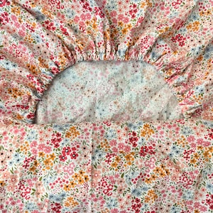 May include: A white fitted sheet with a floral pattern featuring pink, red, orange, and blue flowers. The sheet is made of a soft, comfortable fabric.
