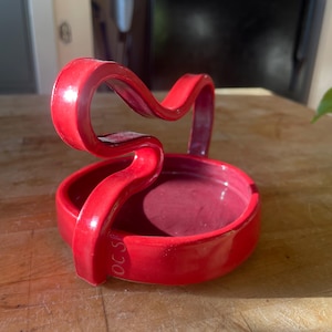 Puede incluir: Un reposacucharas de cerámica rojo vibrante con un diseño de asa curvada único. El reposacucharas tiene una base circular y un acabado brillante. El texto "OC S" es visible en el lateral del asa. Este artículo de cocina está diseñado para sujetar utensilios.