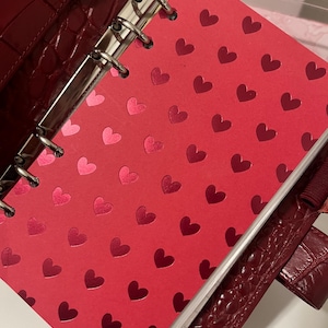 Op de afbeelding: Rode planner met een hartjespatroon. De planner is open en toont een pagina met een herhalend patroon van harten in verschillende tinten rood. De kaft van de planner is dieprood met een gestructureerd, krokodilachtig patroon.