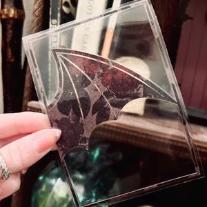 May include: A clear, rectangular sticker with a bat wing design. The wing is filled with a dark floral pattern in shades of burgundy and black. The sticker is held up against a blurred background.