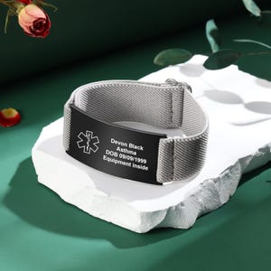 May include: A gray medical alert bracelet with a black plate. The plate has the medical symbol, the name "Devon Black", medical condition "Asthma", date of birth, and the text "Equipment inside".