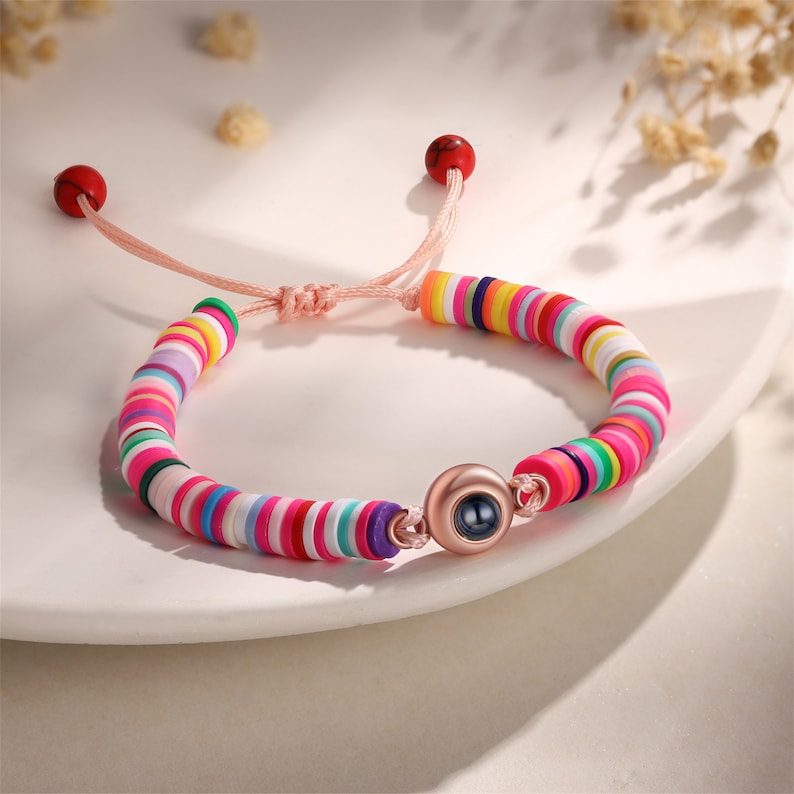 Op de afbeelding: Een kleurrijke kralen armband met een ros&eacute;gouden bedel met een zwart oog. De armband heeft een roze koordsluiting met rode kralen aan de uiteinden.
