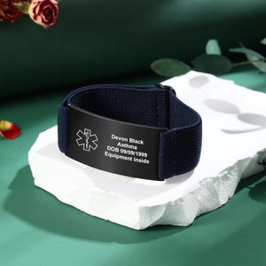 May include: A navy blue medical alert bracelet with a black plate. The plate has a medical symbol and the text "Devon Black, Asthma, DOB 09/09/1999, Equipment inside." The bracelet is on a white surface.