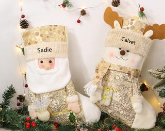 Op de afbeelding: Twee feestelijke kerstsokken, één met een kerstman-thema en één met een rendier-thema. De kerstmansok is goudkleurig met pailletten en de naam "Sadie". De rendiersok is wit en goudkleurig met de naam "Calvet".