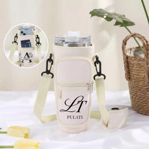 May include: Off-white insulated water bottle carrier with a shoulder strap and a small front pocket. The pocket has a zipper and the initials "LT" and the name "PULATI" printed on it. The carrier also has a small pocket for a phone and cards.