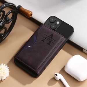 May include: A dark purple leather phone wallet with a slot for cards and a personalized initial "A" with the name "Alexandre" engraved on it. The wallet is attached to a black iPhone.