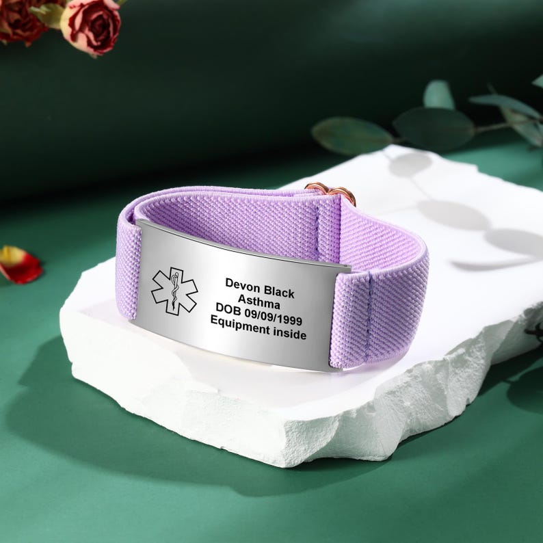 May include: A lavender medical alert bracelet with a silver plate. The plate has a medical symbol and the text: "Devon Black, Asthma, DOB 09/09/1999, Equipment inside." The bracelet is on a white surface.
