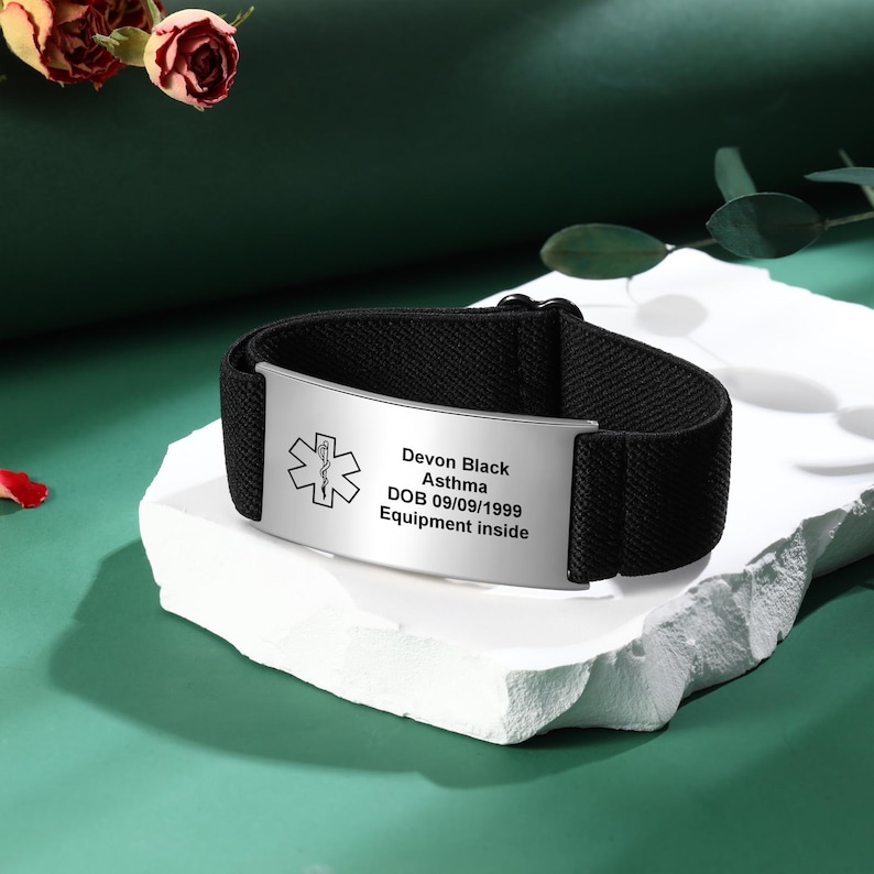 May include: Black medical alert bracelet with a silver metal plate. The plate has a medical symbol and the text: Devon Black, Asthma, DOB 09/09/1999, Equipment inside. The bracelet is on a white surface.