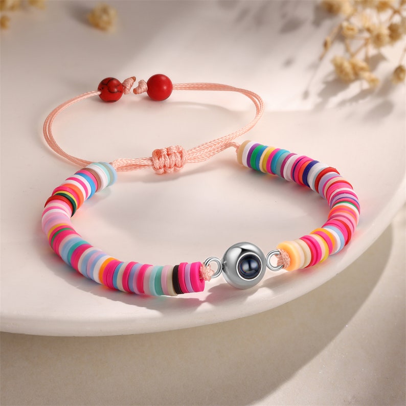 Op de afbeelding: Een kleurrijke kralen armband met een zilveren bedel met een kleine zwarte parel. De armband is gemaakt met een roze koord en heeft rode kralen aan de uiteinden.