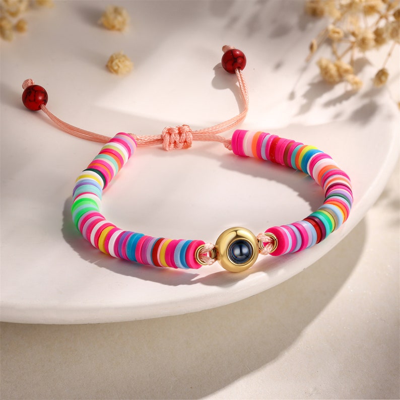 Op de afbeelding: Een kleurrijke kralen armband met een roze koord en een gouden bedel met een zwart oog. De kralen zijn gerangschikt in een regenboogpatroon.