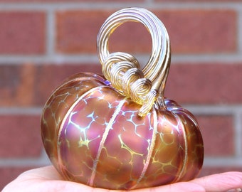 May include: A glass pumpkin with a swirling gold stem. The pumpkin is iridescent with shades of orange, pink, and gold.