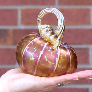 May include: A glass pumpkin with a swirling gold stem. The pumpkin is iridescent with shades of orange, pink, and gold.