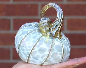 May include: A white glass pumpkin with a gold swirl design. The pumpkin has a textured surface and a gold stem.