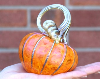 Puede incluir: Una calabaza de vidrio con un diseño de espiral en naranja y dorado. La calabaza tiene un acabado brillante y metálico.