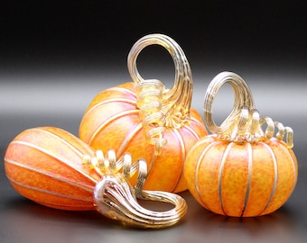 May include: Three glass pumpkins in orange and gold with a swirl design. The pumpkins are arranged in a row with the largest pumpkin in the back and the smallest pumpkin in the front.