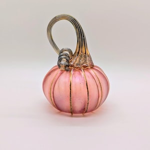 May include: A pink glass pumpkin with gold accents and a curved stem. The pumpkin has vertical gold stripes and a looped stem. The glass is translucent, allowing light to pass through. A decorative fall-themed item.