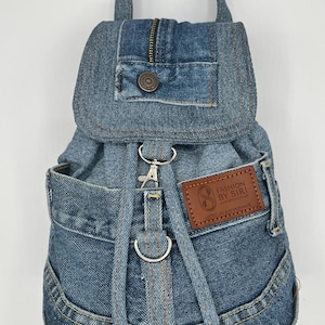 May include: A small, blue denim backpack with a top handle and adjustable straps. The backpack features a front flap with a zipper and a button closure. A brown leather patch with the text "FASHION BY SIRI" is sewn on the front. The backpack is made from recycled materials.