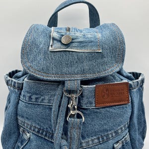 May include: A blue denim backpack with a top handle and adjustable straps. The bag features a flap closure with a button and a small brown leather patch with the text "FASHION BY SIRI". The backpack has silver-tone hardware and a drawstring closure.
