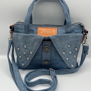May include: A blue denim handbag featuring two top handles and a detachable shoulder strap. The front displays two triangular flaps adorned with silver-tone metal studs, a central vertical placket with two metal buttons, and a tan leather patch.