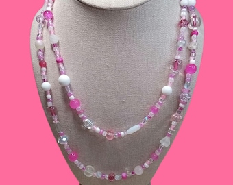 May include: A layered necklace featuring a variety of pink, white, and clear beads. The beads vary in size and shape, including round, oval, and faceted designs. The necklace is displayed on a mannequin with a pink background.