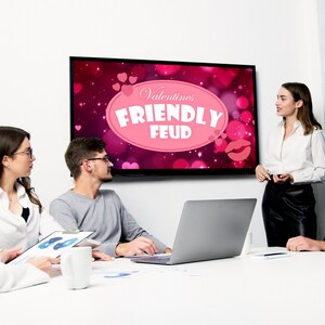 May include: A group of people are gathered around a table, looking at a screen that displays the words "Valentine's Friendly Feud". The screen has a pink background with hearts and a pink oval with the text.
