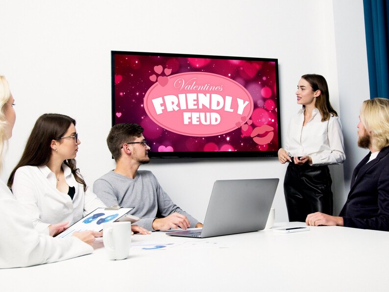 May include: A group of people are gathered around a table, looking at a screen that displays the words "Valentine's Friendly Feud". The screen has a pink background with hearts and a pink oval with the text.