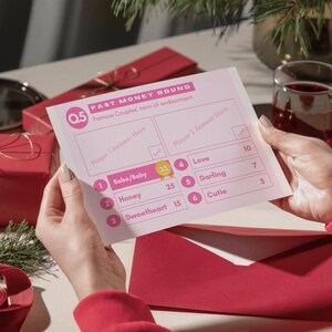 May include: A pink and white game card with the title "Fast Money Round" and the text "Famous Couples' terms of endearment." The card has a list of terms of endearment with points assigned to each term. The card is being held by a person's hands.