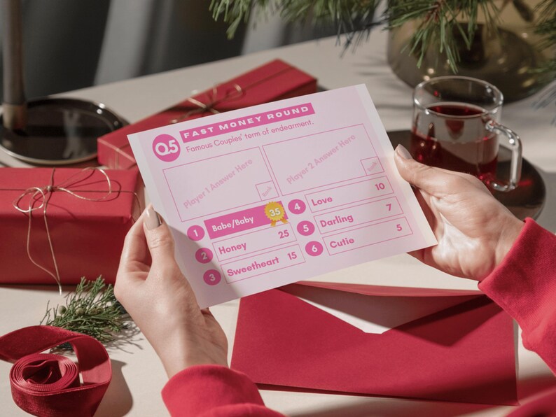 May include: A pink and white game card with the title "Fast Money Round" and the text "Famous Couples' terms of endearment." The card has a list of terms of endearment with points assigned to each term. The card is being held by a person's hands.