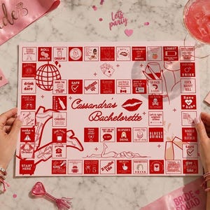 May include: A pink and red bachelorette party game board with the text "Cassandra's Bachelorette" and various game instructions. Includes a pink sash, dice, and a cocktail.