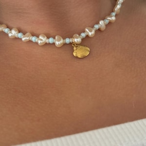 Op de afbeelding: Een delicate choker ketting gemaakt met kleine, crèmekleurige parels en een lichtblauwe tussenparels. De ketting heeft een gouden schelp hanger.