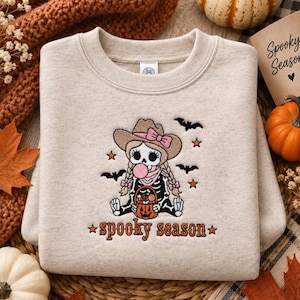 Puede incluir: Una sudadera beige de cuello redondo con un diseño bordado de un esqueleto con sombrero de vaquero y trenzas, haciendo una burbuja rosa. El esqueleto sostiene un cubo de calabaza lleno de dulces, rodeado de murciélagos y estrellas, con el texto "spooky season" debajo.