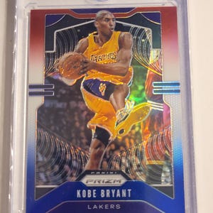 May include: A basketball trading card featuring Kobe Bryant in a yellow Lakers uniform, holding a basketball. The card has a blue and red border and the text "Kobe Bryant" and "Lakers". The card is encased in a clear plastic holder.