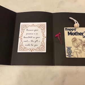 Op de afbeelding: Een zwarte Moederdagkaart met een decoratief label en een klein bloemstuk. Op het label staat "Happy Mother's Day" en het bevat een illustratie van een moeder en kind. Een kleinere kaart binnenin heeft de tekst "Because your presence is as beautiful as your scent... this gift is made for you."