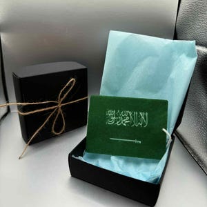 Op de afbeelding: Een zwarte geschenkdoos vastgebonden met touw naast een open zwarte doos met een groene kaart erin. De groene kaart toont witte Arabische tekst en een wit zwaard op een groene achtergrond, op blauw vloeipapier.