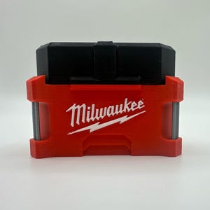 Puede incluir: Un accesorio de herramienta rojo y negro con el logotipo de Milwaukee en blanco. El accesorio tiene forma rectangular con una parte superior negra y una base roja. El logotipo de Milwaukee está centrado en la base roja.