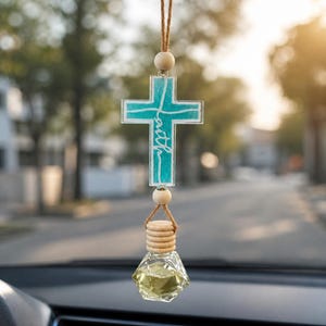 May include: A car air freshener with a teal cross pendant that says "faith" in cursive. The cross is suspended by a brown string and wooden beads. Below the cross is a glass bottle with a yellow liquid.