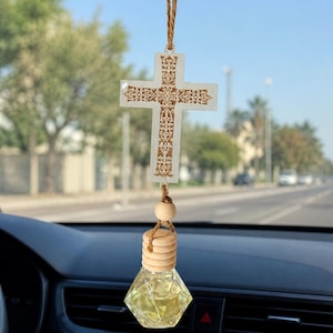 May include: Car air freshener with a decorative cross pendant. The cross is white with a brown, ornate design. Below the cross hangs a small, clear, geometric-shaped bottle filled with yellow liquid, topped with a wooden cap.