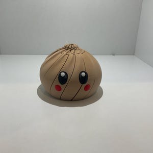 May include: A tan, rounded, 3D-printed dumpling figurine featuring a spiral-textured top, two large black eyes with white highlights, and two small red circular cheeks. The object has a smooth, matte finish and a symmetrical, bulbous shape.