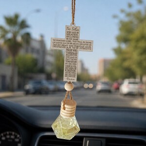 May include: A car air freshener with a wooden cross pendant featuring the Lord's Prayer. The cross is suspended from a brown string, with a wooden bead and a glass bottle containing a yellow liquid. The background shows a blurred view of a road.