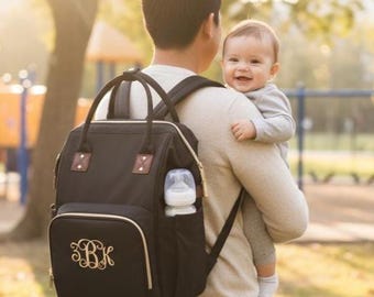 May include: Black diaper bag with gold trim, a front pocket with embroidered initials, and a side pocket holding a baby bottle. The bag has a top handle and adjustable backpack straps. A person is carrying a baby in a park setting.