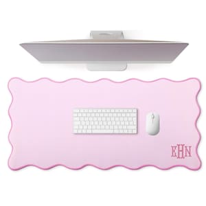 Puede incluir: Alfombrilla de escritorio rosa claro con borde festoneado, teclado blanco y ratón blanco. La alfombrilla tiene un monograma en la esquina inferior derecha y un monitor de ordenador en el fondo.