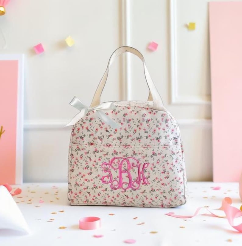 Może przedstawiać: Torba na lunch z kwiatowym nadrukiem z białym uchwytem i r&oacute;żowym monogramem. Torba ma kremowe tło ze wzorem małych r&oacute;żowych kwiat&oacute;w i zielonych liści. Biała kokarda jest zawiązana w pobliżu uchwytu.