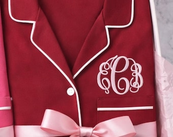 May include: A deep red button-down pajama shirt featuring white piping along the collar, front placket, and chest pocket. A white embroidered monogram is centered on the pocket. The shirt is wrapped with a light pink satin ribbon in a box.