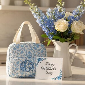 Może przedstawiać: Torba na lunch z kwiatowym nadrukiem i monogramem, bukiet niebieskich ostr&oacute;żek i białych r&oacute;ż w białym dzbanku oraz kartka z napisem "Happy Mother's Day". Torba na lunch ma jasnoniebieski kwiatowy wz&oacute;r.